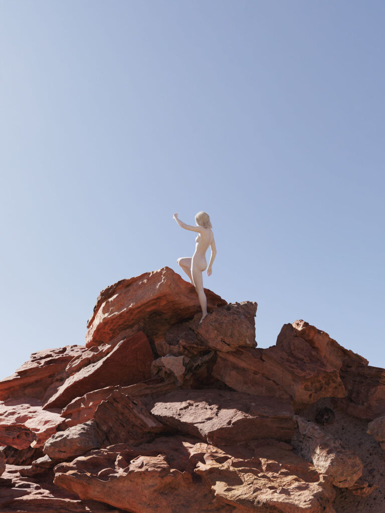 A woman with blonde hair stands atop rugged red rock formations under a clear blue sky reaching out with one hand.